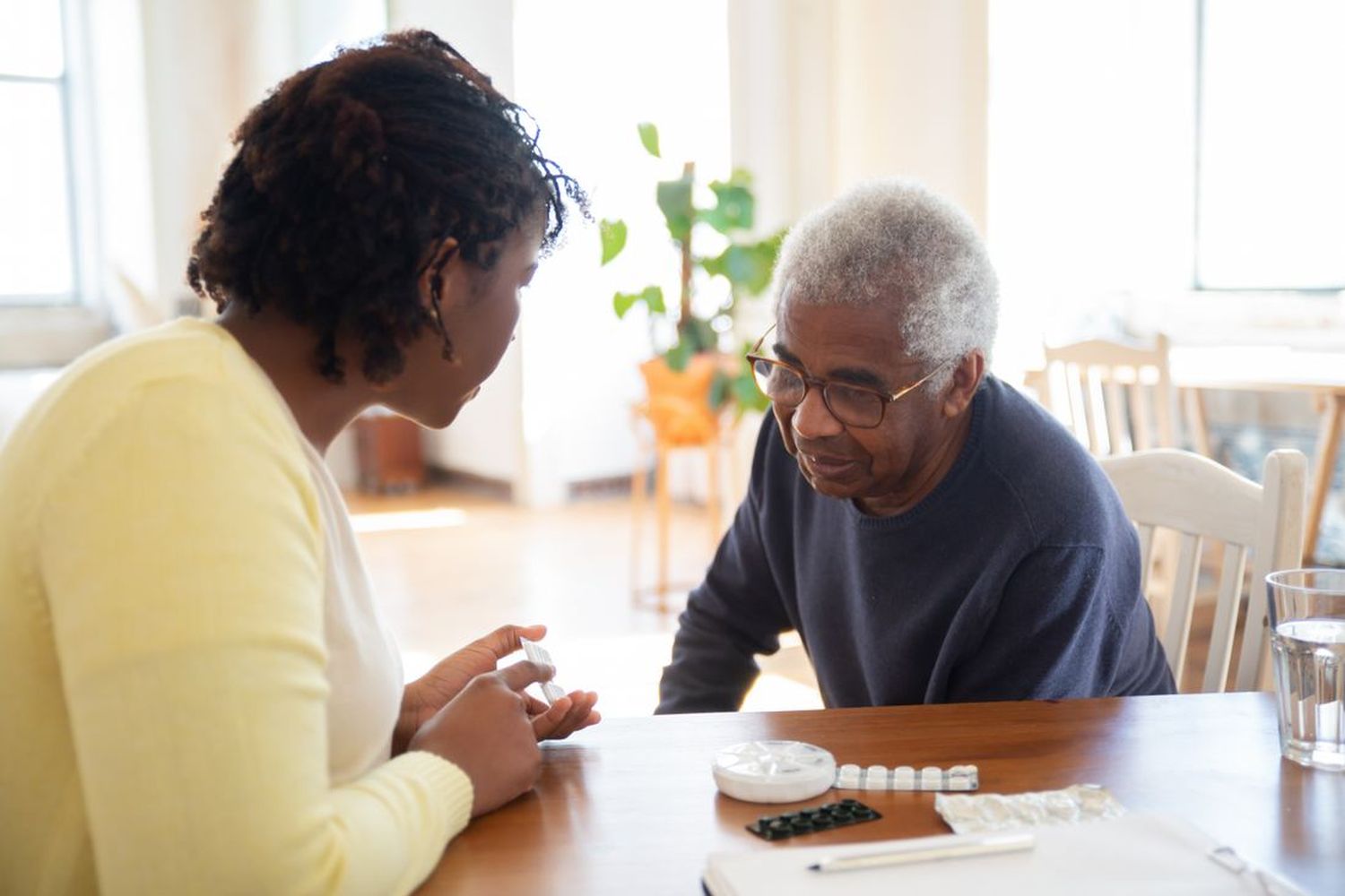 A woman helping an older man sort his medication at a sunlit kitchen table